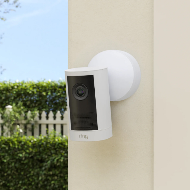 Ring Outdoor Camera 4K Plus on a beige wall overlooking a back garden with a white fence and hedges.