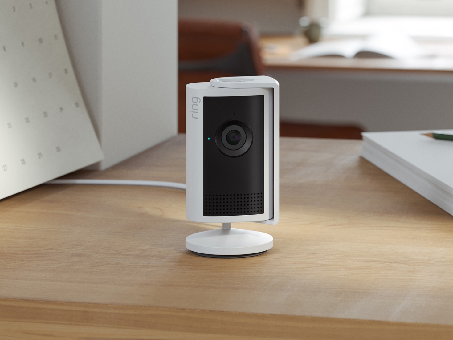 White Indoor Camera Plus sits on a surface indoors next to decorative books and a potted plant. 