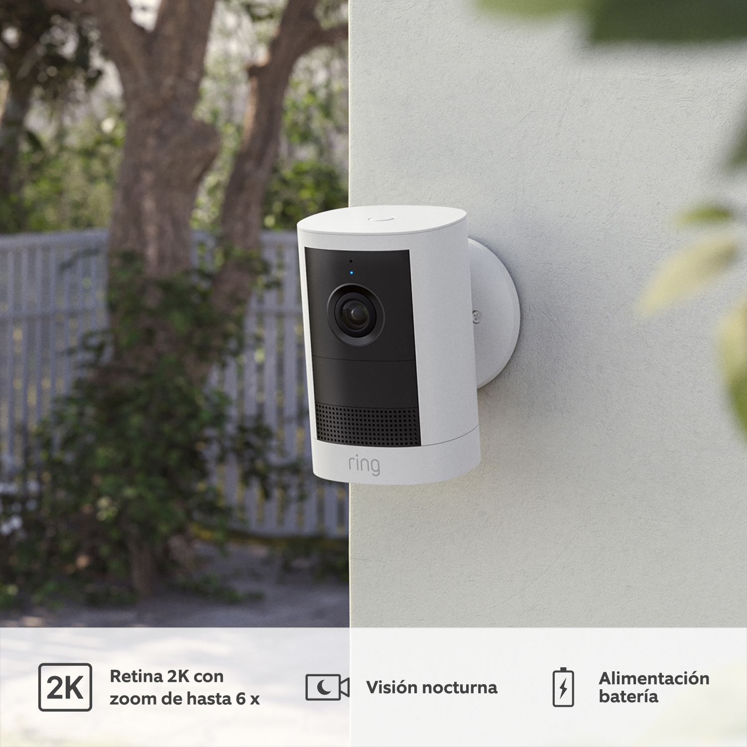 Cámara exterior con batería (última generación) (for Outdoor Camera Battery) - Ring Outdoor Camera (2nd Gen) blanca en pared exterior gris, con lente y rejilla negras. Follaje al fondo.