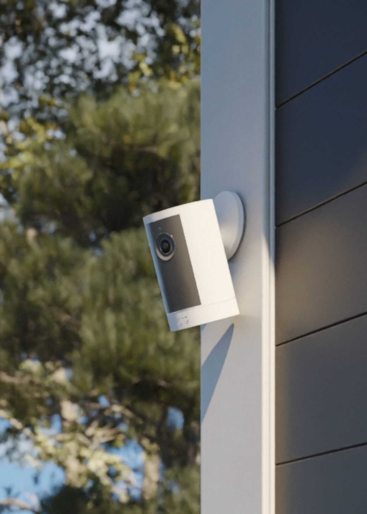 Ring Outdoor Camera 4K Plus in white mounted on the corner of a home, overlooking a tree-lined outdoor area.
