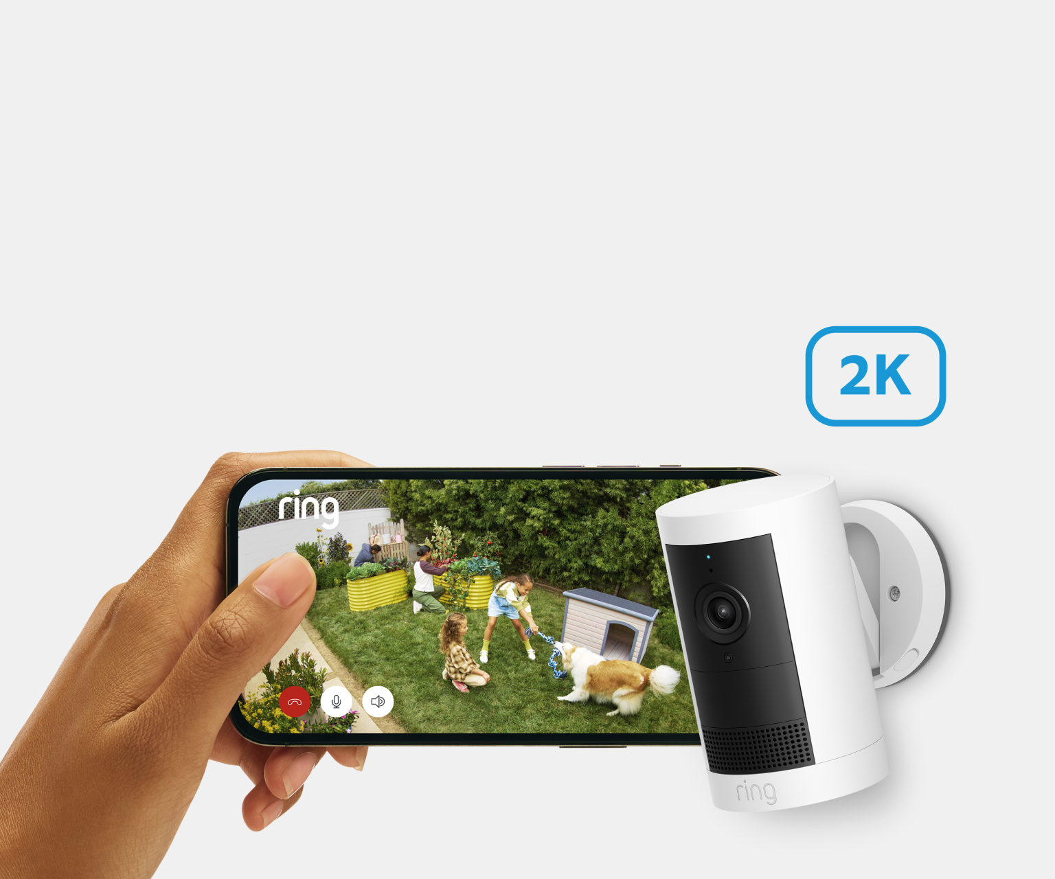 Person holding phone showing Ring 2K camera footage of children and dog playing in garden, next to wall-mounted camera.