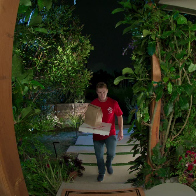Delivery person in uniform is on a front path holding branded pizza box and paper bag. Scene is well-lit with vibrant colours