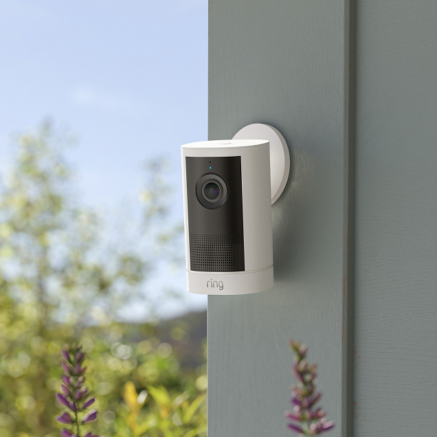 Ring Outdoor Camera 4K Plus mounted on the side of a house, angled toward the front garden with trees behind.
