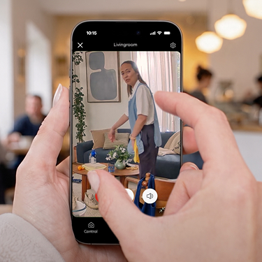 Manos con smartphone mostrando Live View de Pan-Tilt Indoor Camera 2K con mujer en delantal azul en casa.
