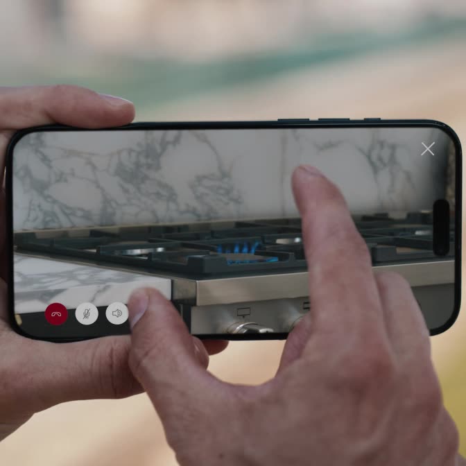 Hands holding a horizontal phone, reverse pinching to zoom in on a stainless steel gas stove with a lit burner with blue flames.
