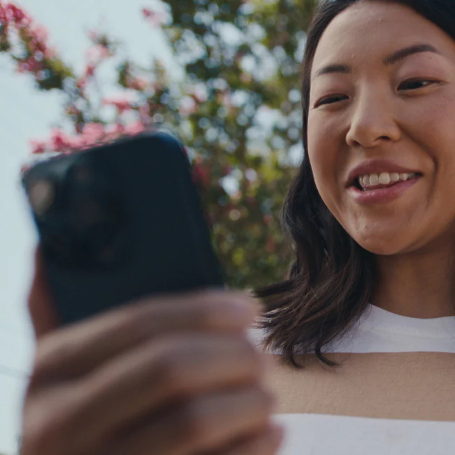 Femme souriante regarde son smartphone en extérieur, interagissant probablement avec l'application Ring.