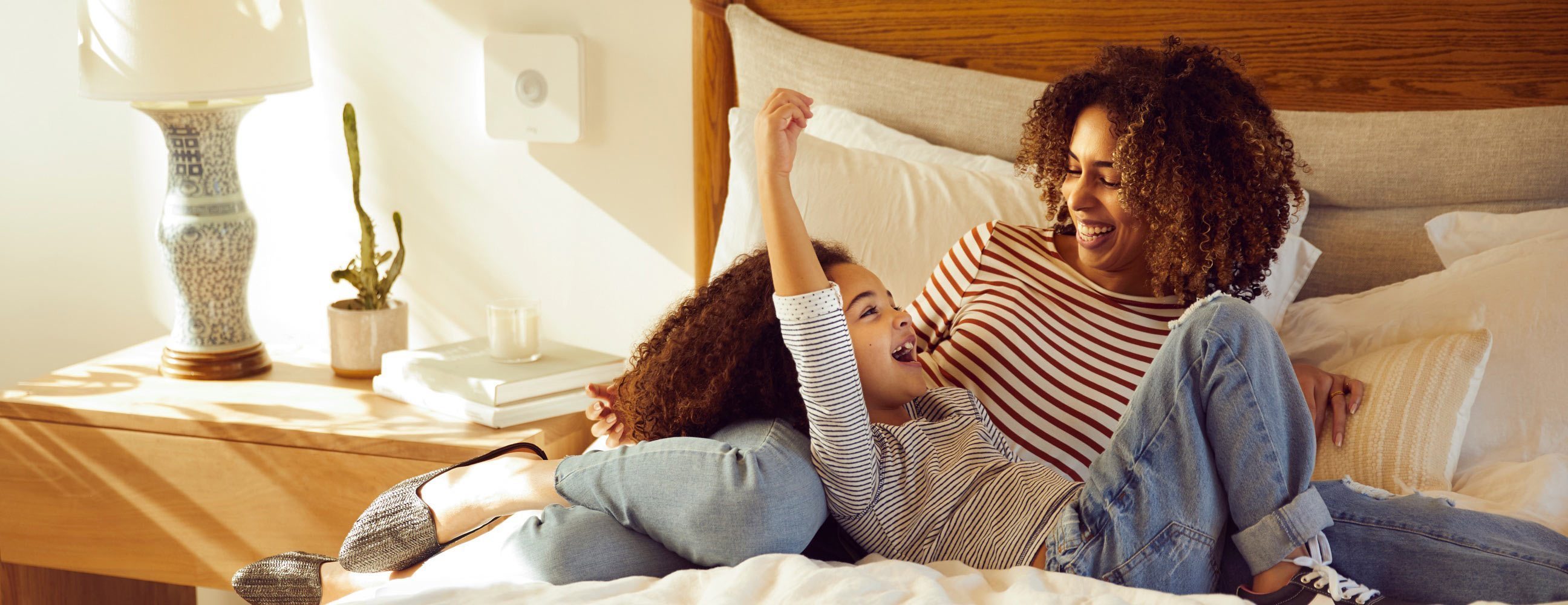 Mutter und Tochter lachen zusammen auf dem Bett, Ring Base Station sichtbar an der Wand über dem Nachttisch.