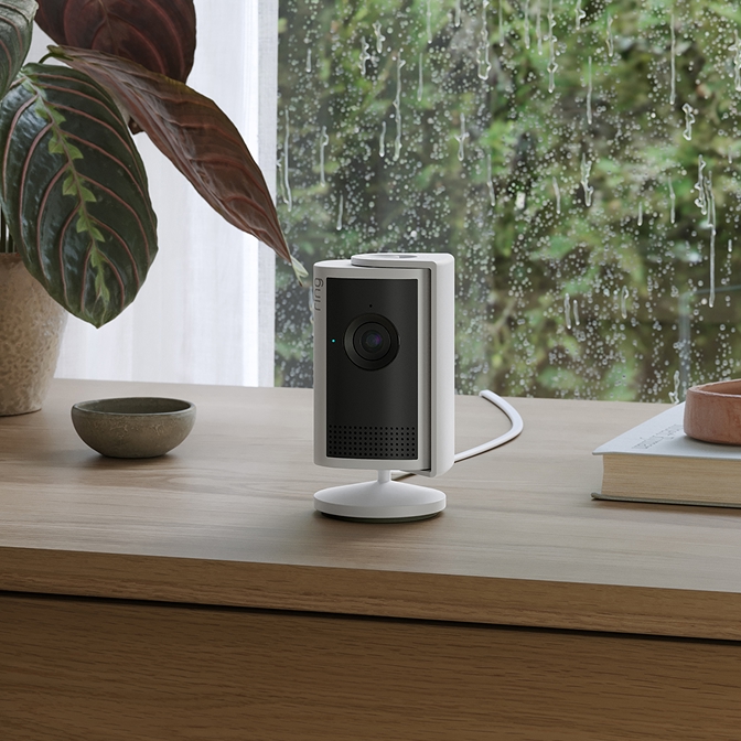 Een witte Indoor Camera Plus staat binnen op een vlakke ondergrond, naast decoratieve boeken en een bloempot.