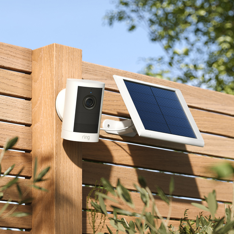 Outdoor Camera (2nd Gen) is installed on a brown outdoor fence with solar panel attached beside and foliage in the background