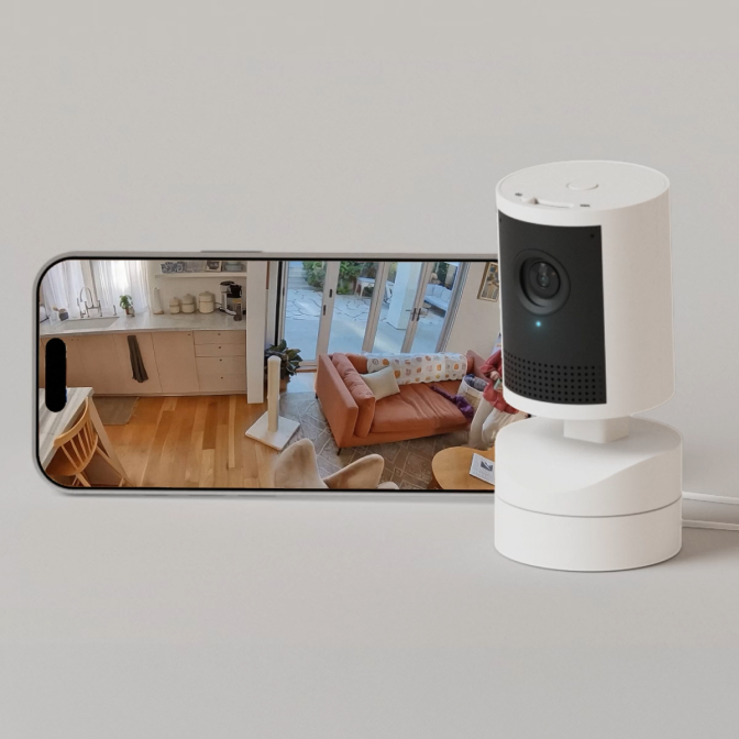 Cámara Pan-Tilt Indoor Camera 2K blanca en estante junto a smartphone mostrando vista en vivo con controles pan-tilt.