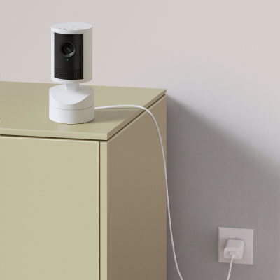 Cámara interior Pan-Tilt 2K blanca en esquina de mueble con cable conectado a enchufe de pared.