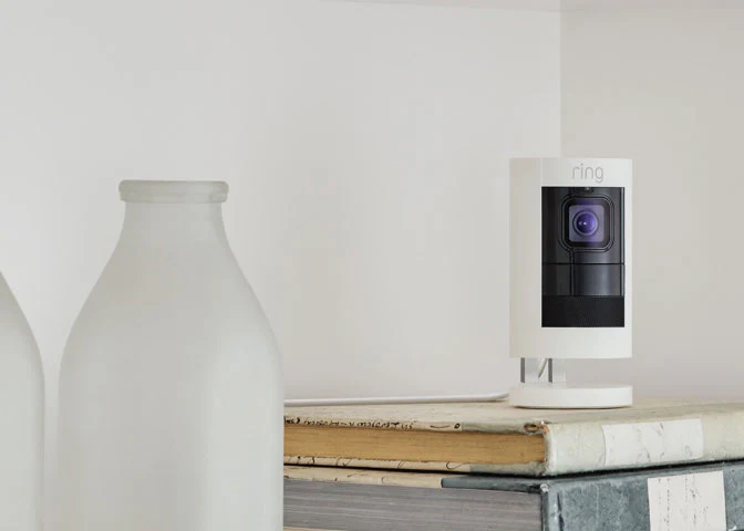 White Ring Stick Up Cam Elite placed on stacked books indoors near frosted glass bottle.