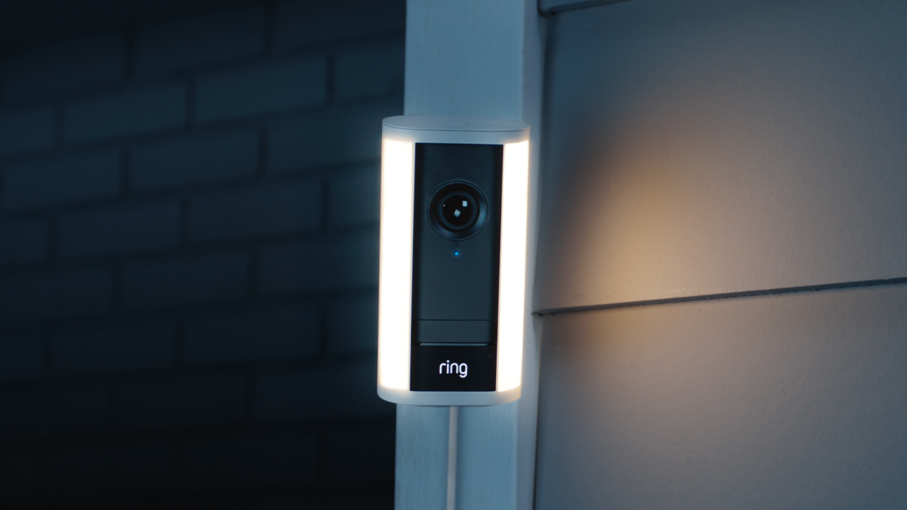 A wall-mounted Ring Spotlight Cam Pro activates suddenly at dusk, phones shows intruder in garden via Ring app. User taps siren button, causing intruder to flee