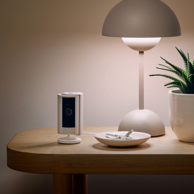 Ring Indoor Camera on a wooden table near a lamp, plant, and keys in a softly lit room.