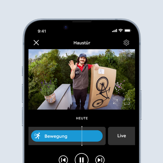 Smartphone zeigt Live-Video der Ring App von Paketboten mit Paket, der an der Haustür winkt.