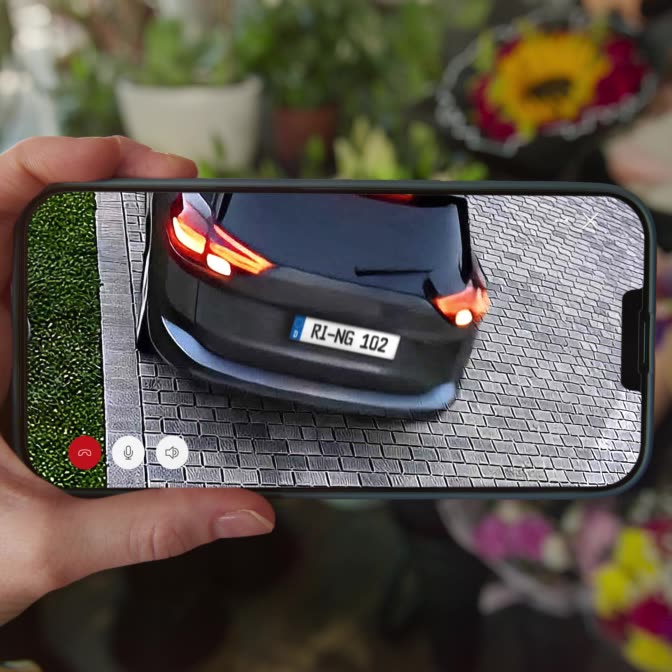 Two hands holding a phone with the Ring app on the screen. The fingers of one hand pinch and expand the view to zoom in on the number plate of a car which is driving away from the property.