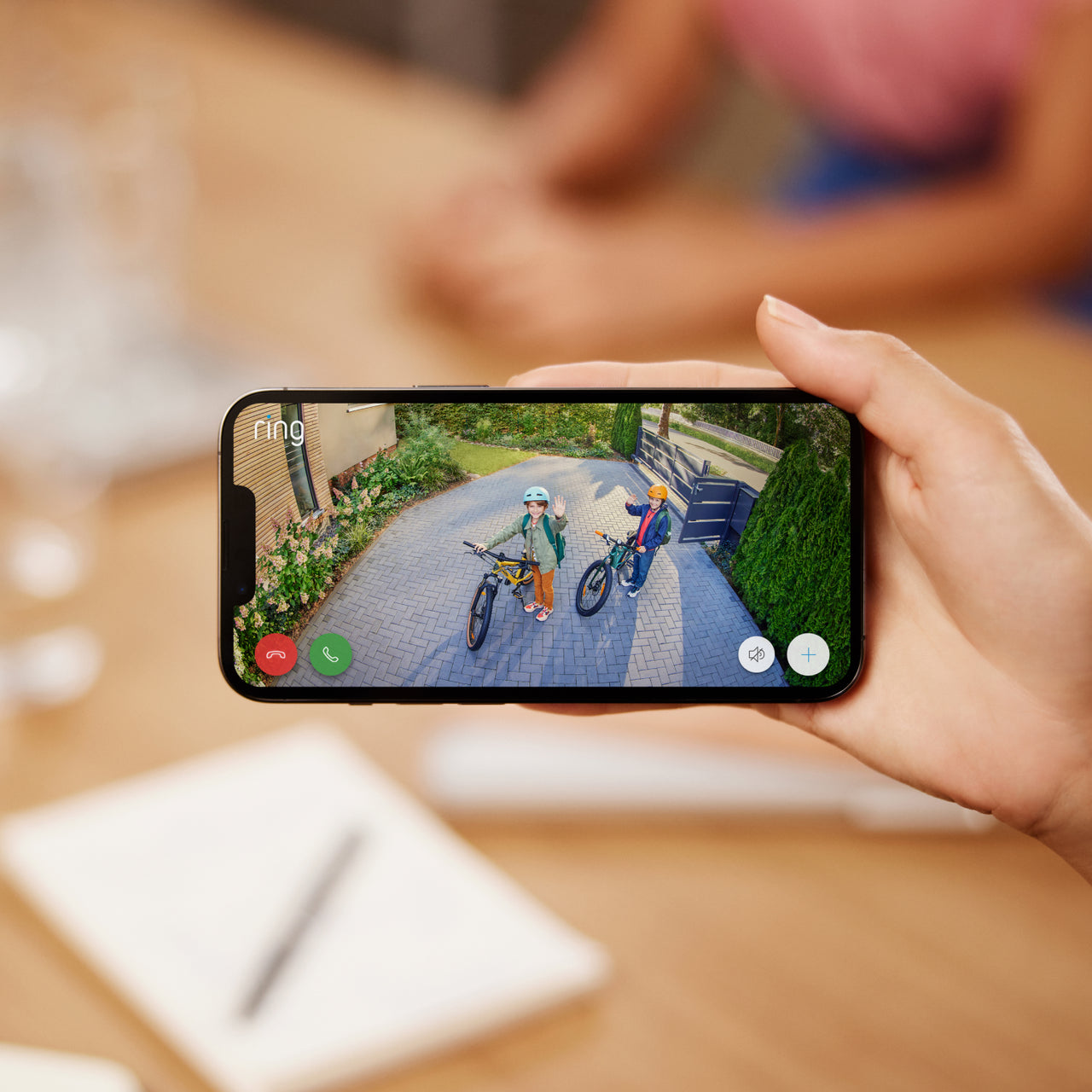 3-Pack Stick Up Cam Elite - Person using the Ring app on a smartphone to view and talk to two children outside riding bikes.