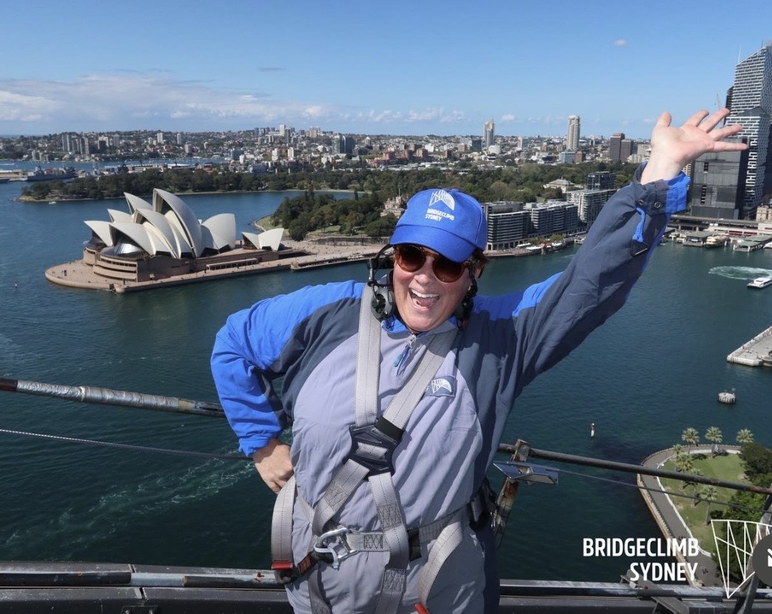 A picture of Audrey Weber on a bridge overlooking the Sydney Opera House. She's looking at the camera and waving.