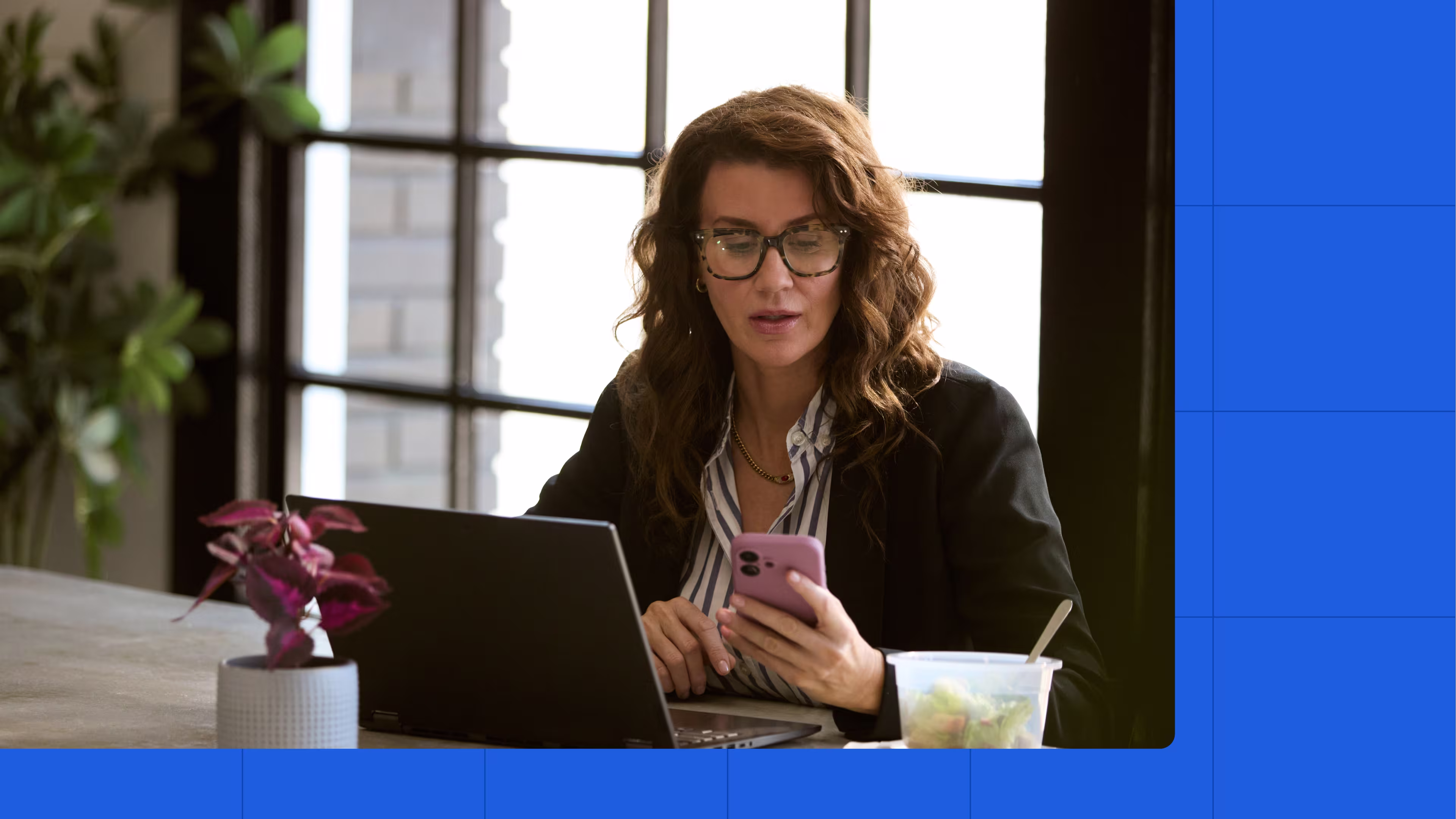 A woman sitting at a table, looking at her phone while working on her laptop.