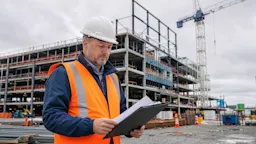 A construction site supervisor in a bright orange high-visibility vest and white hard hat reviewing a clipboard of legal documents at a large commercial construction site in Ontario, Canada.