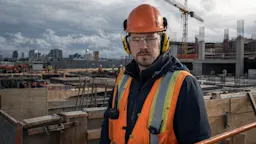 A construction worker wearing yellow earmuffs and an orange high-visibility vest stands on a commercial construction site in Canada.