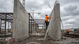 A Canadian construction worker in a bright orange high-visibility vest and white hard hat climbing a tall aluminum extension ladder leaning against a concrete wall at an active construction site.