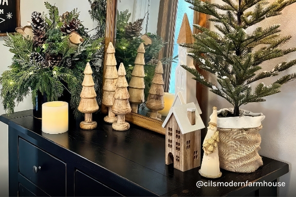 Instagram handle @cilsmodernfarmhouse. Table with assorted ferns and pinecones, wood trees, mirror, and cream-colored candle.