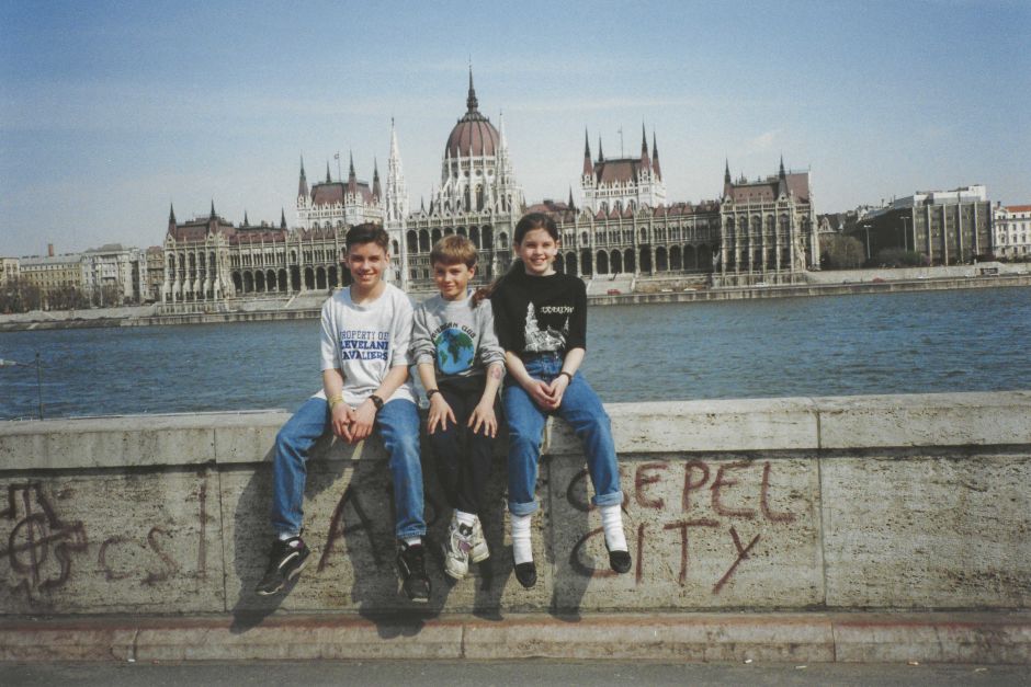 Stephen seated between his elder siblings, Nicholas and Emelia, in 1994. The Danube and the Hungarian Parliament building appear in the background. 