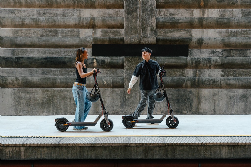 Två personer står med varsin Segway-elsparkcykel framför en betongvägg.