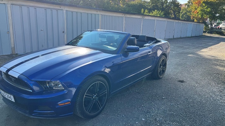 Vista frontal de un Ford Mustang convertible azul con llantas negras y franjas deportivas, estacionado al aire libre.