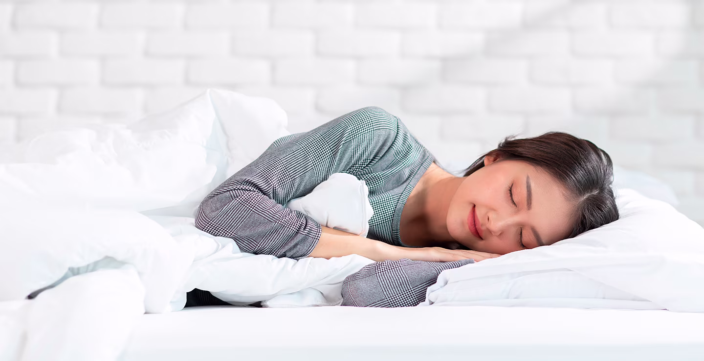 Person lying on a bed with white bedding in a bright bedroom with a white brick wall background.