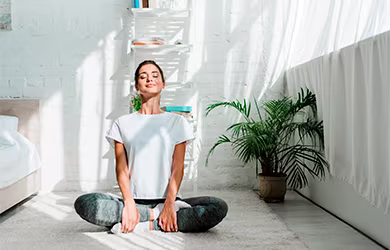 A woman with her eyes closed sitting cross-legged on a white rug in a sunlit room, meditating or practicing yoga.