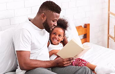 African father reading with his adorable little daughter at home in bed.