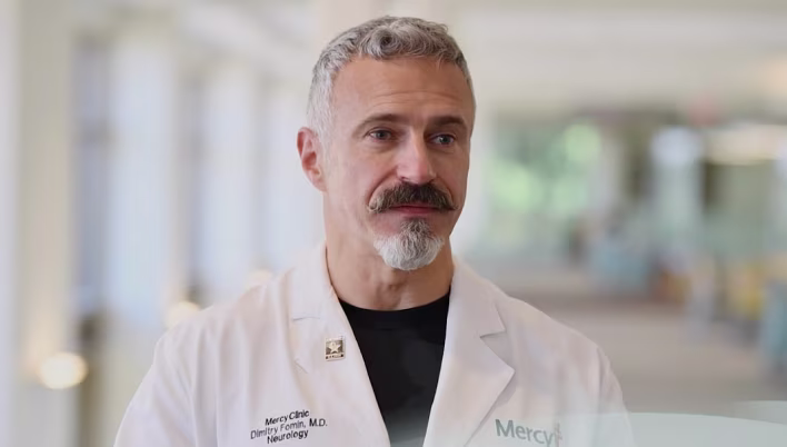 A healthcare professional in a white lab coat with visible text "Mercy Clinic" and "Neurology," standing in a bright indoor setting with blurred background.