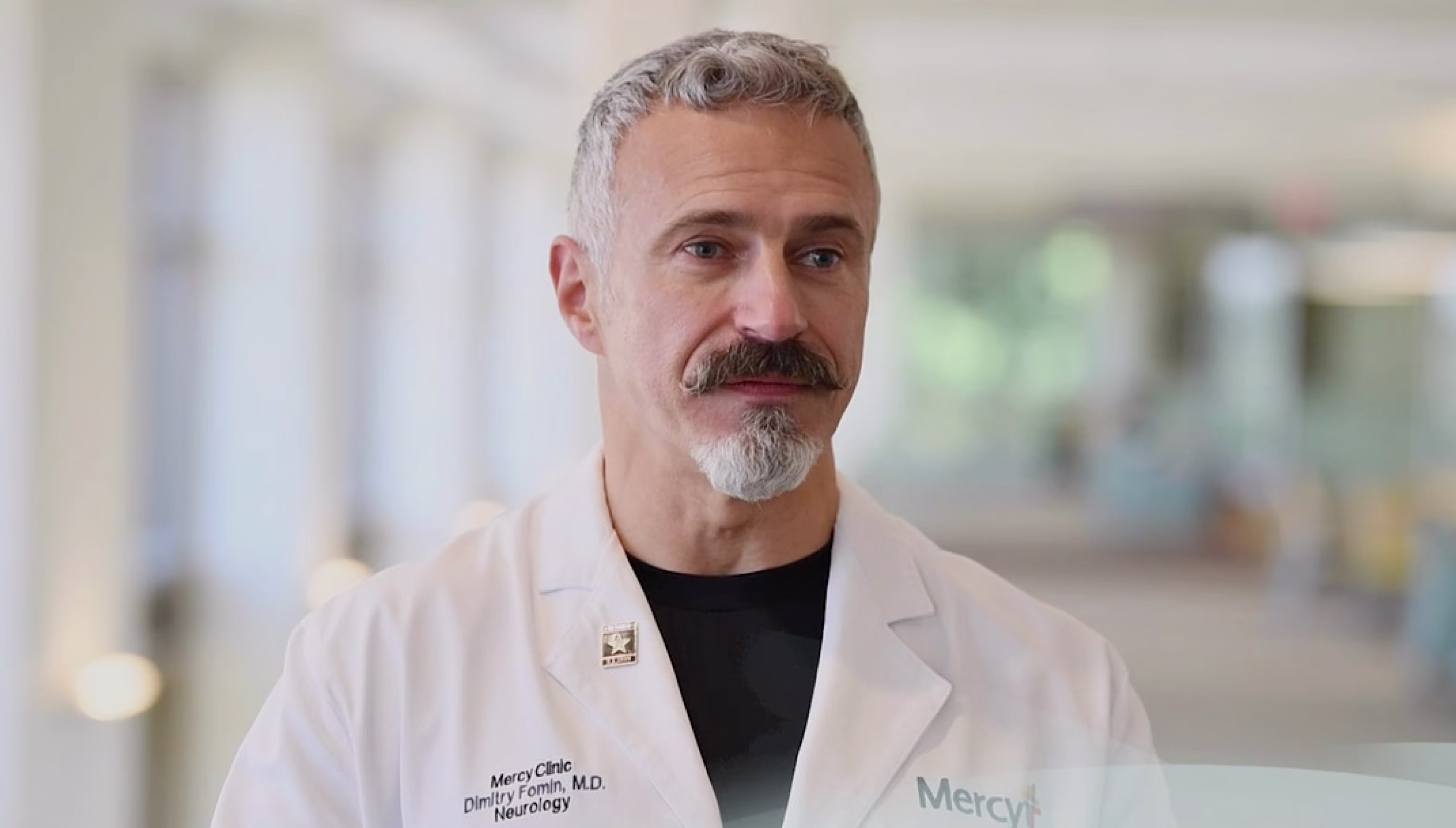 A healthcare professional in a white lab coat with visible text "Mercy Clinic" and "Neurology," standing in a bright indoor setting with blurred background.