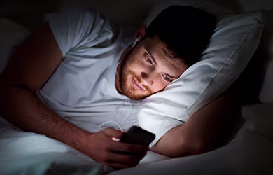 A man lying on a bed, browsing his phone and showing signs of sleep troubles.