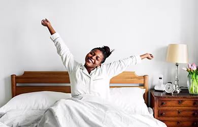 A woman in pajamas stretching arms while sitting on a bed with white bedding and wooden headboard.