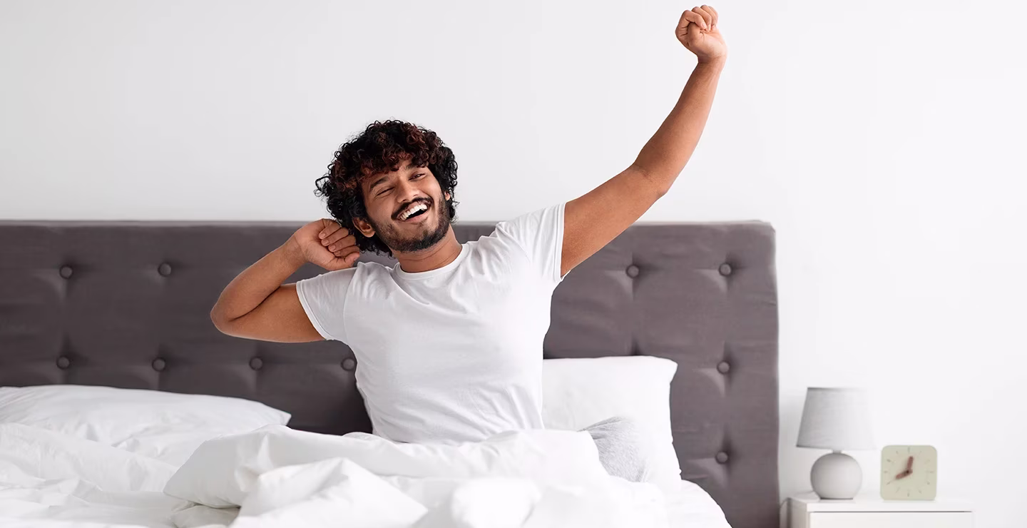 A man is smiling and stretching after waking up, wearing a white t shirt.