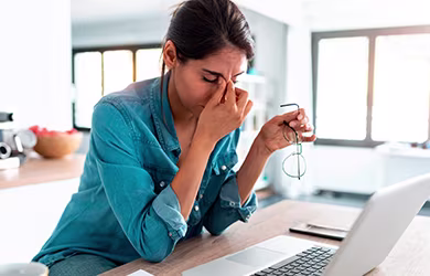 A woman sitting at a table with a laptop, holding her glasses and pinching the bridge of her nose, appearing stressed or experiencing eye strain.