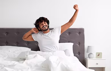 A happy man with curly hair sits up in bed, smiling and stretching his arms after waking up.