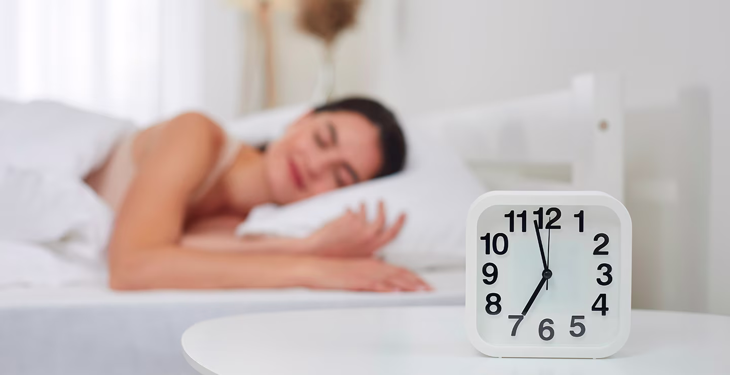 White analog clock showing 7:35 on a bedside table with a person sleeping in the background.