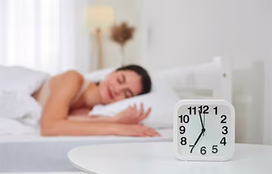 White analog clock showing 7:35 on a bedside table with a person sleeping in the background.