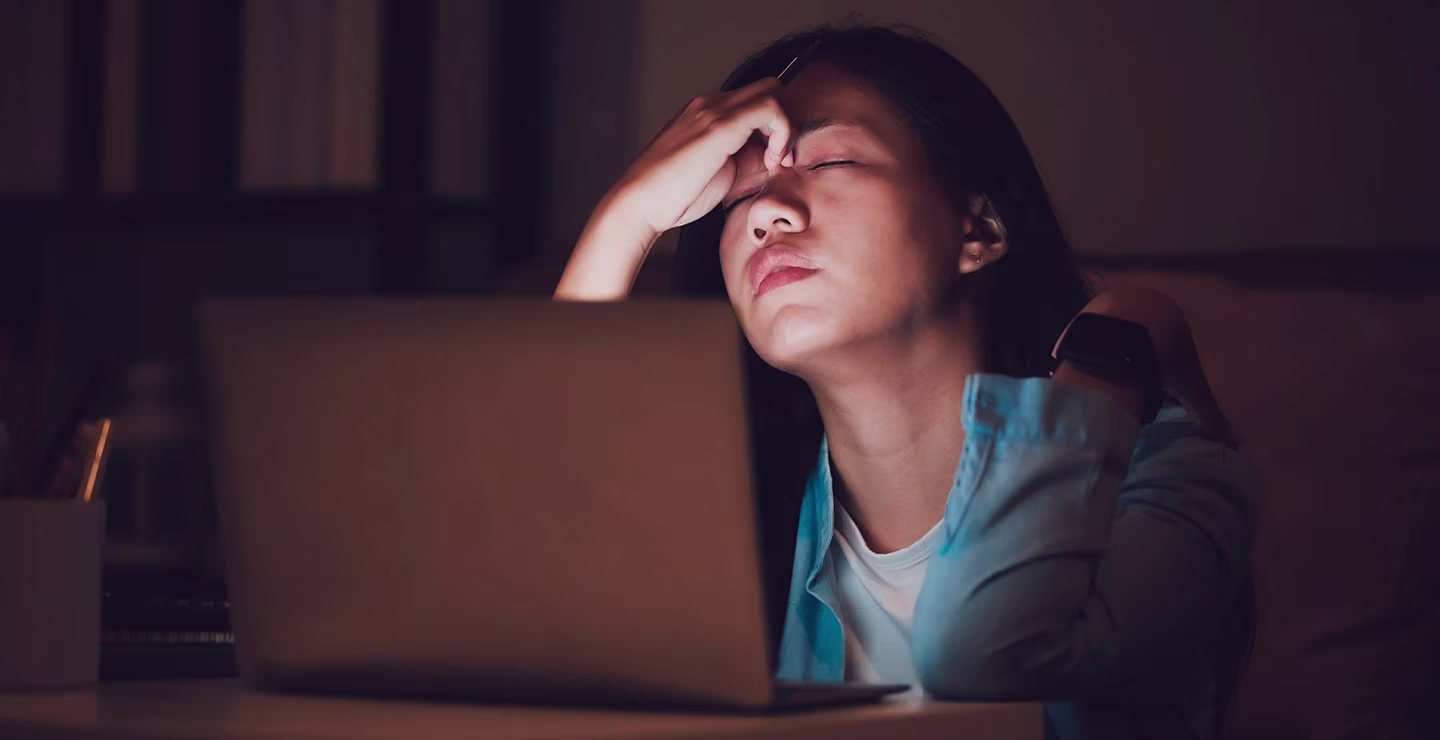 A woman sitting in the dark in front of a laptop, holding her forehead in distress.