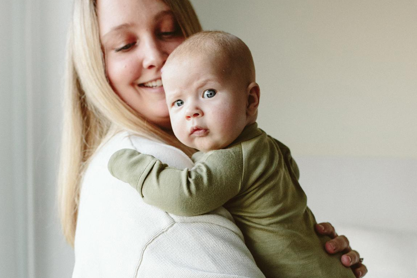 Smiling blonde woman holding a blue-eyed baby wearing an olive green outfit against a neutral background.