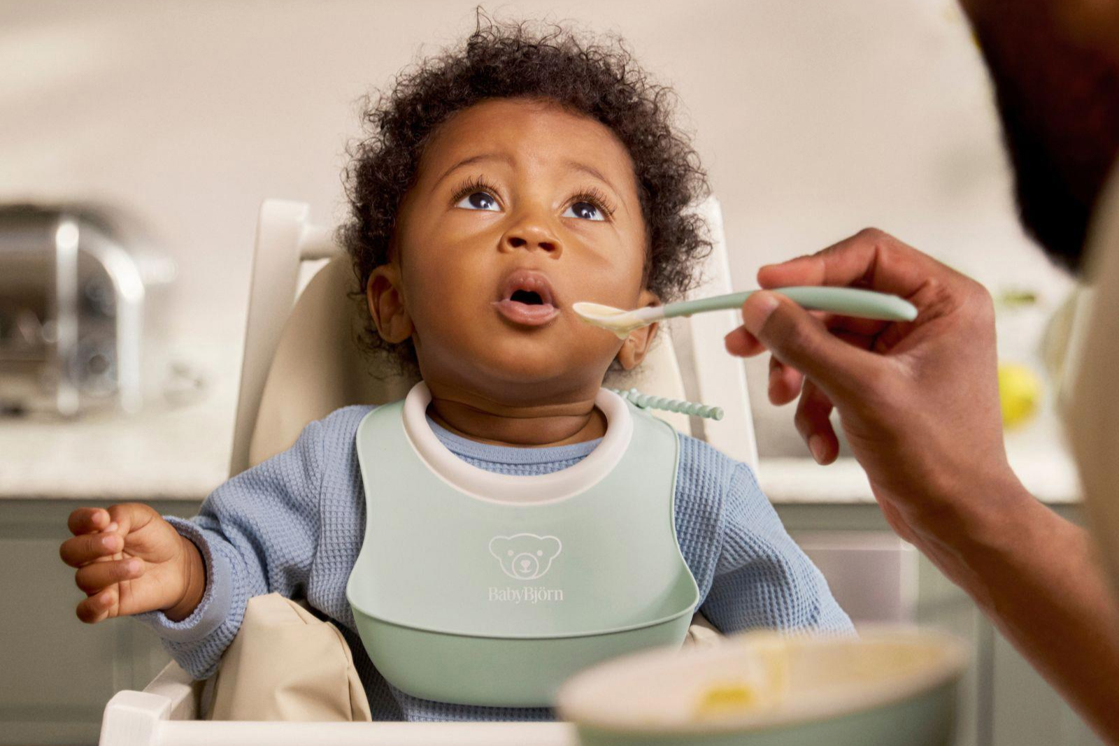 Young child with curly hair wearing a light blue bib being spoon-fed by an adult in a high chair.