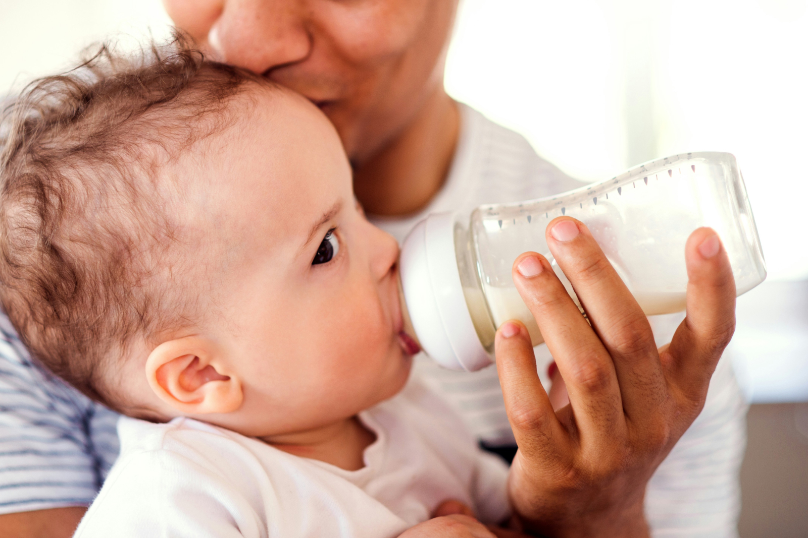 Parent bottle-feeding infant with milk while gently kissing baby's head in a tender moment of care and bonding.