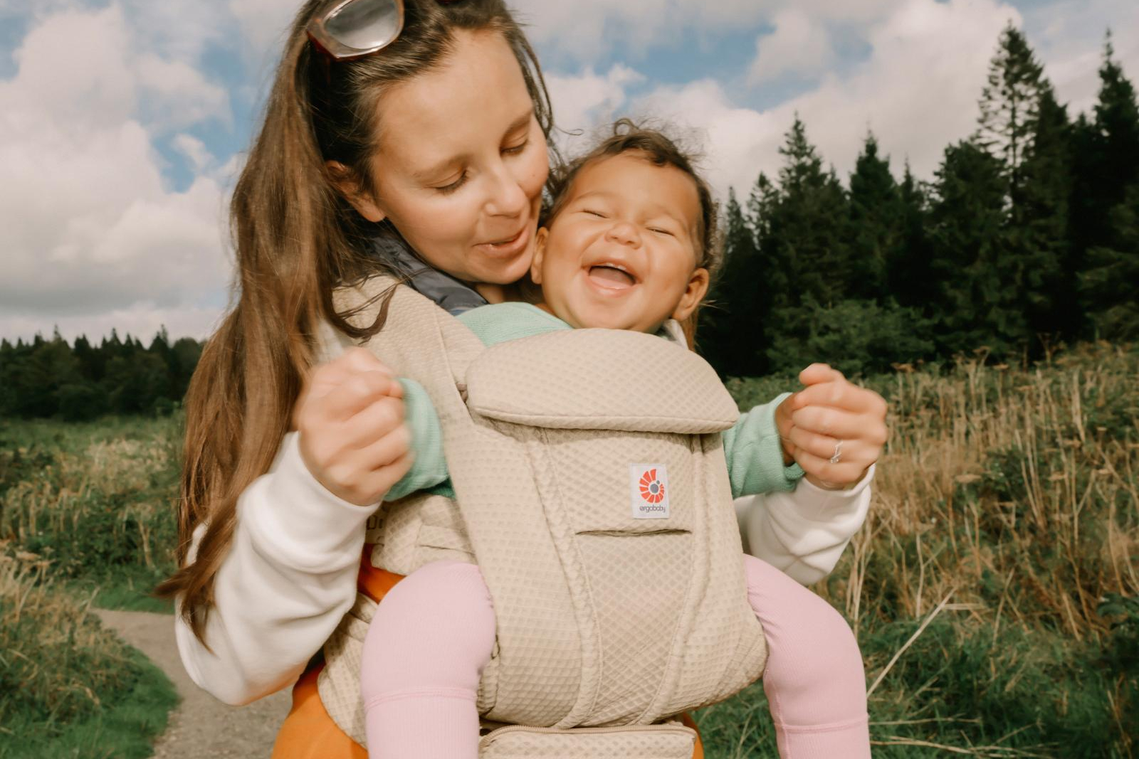 Mother carrying smiling baby in beige carrier outdoors with evergreen trees and cloudy sky in background.