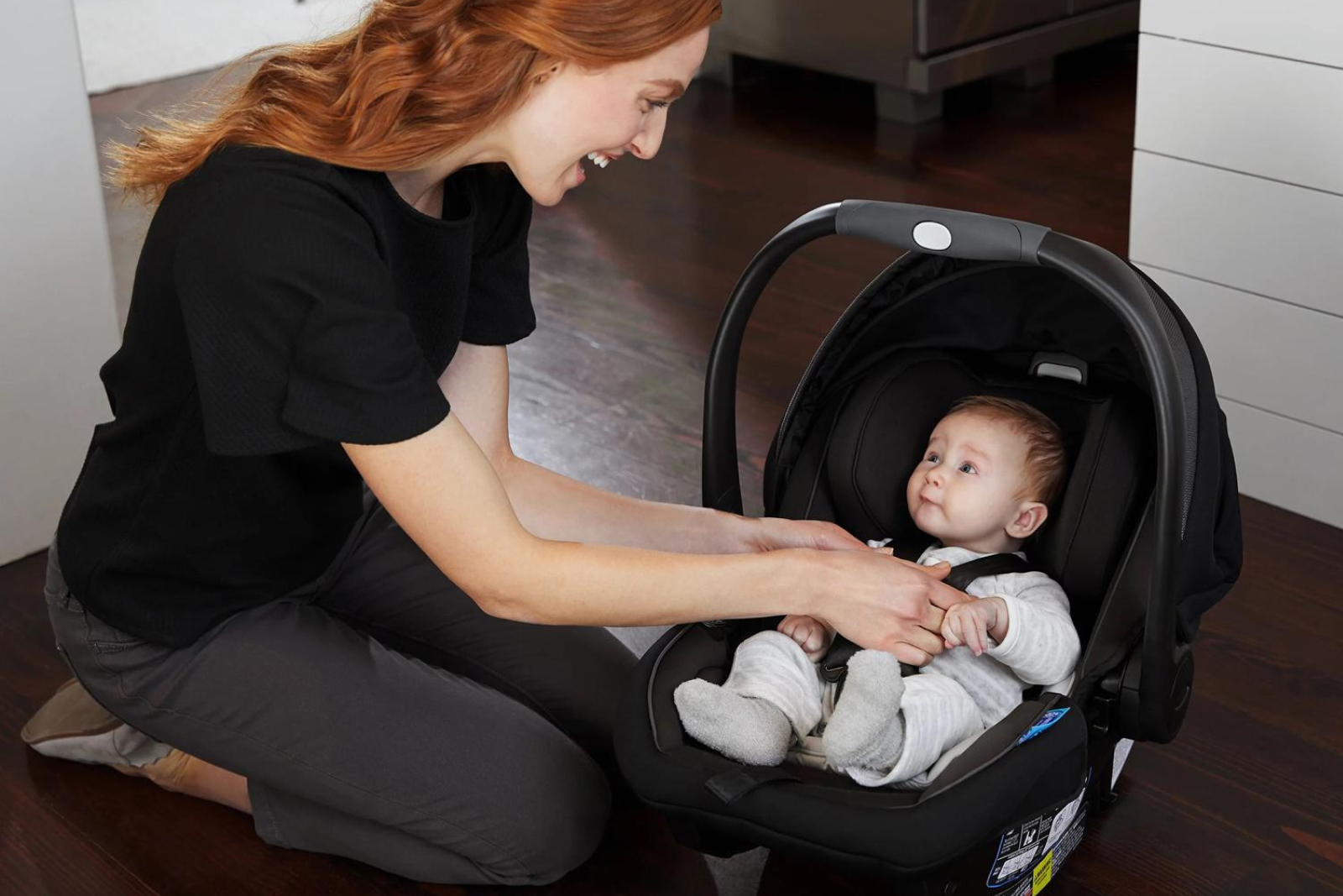 Woman with red hair in black shirt securing baby in black car seat on wooden floor
