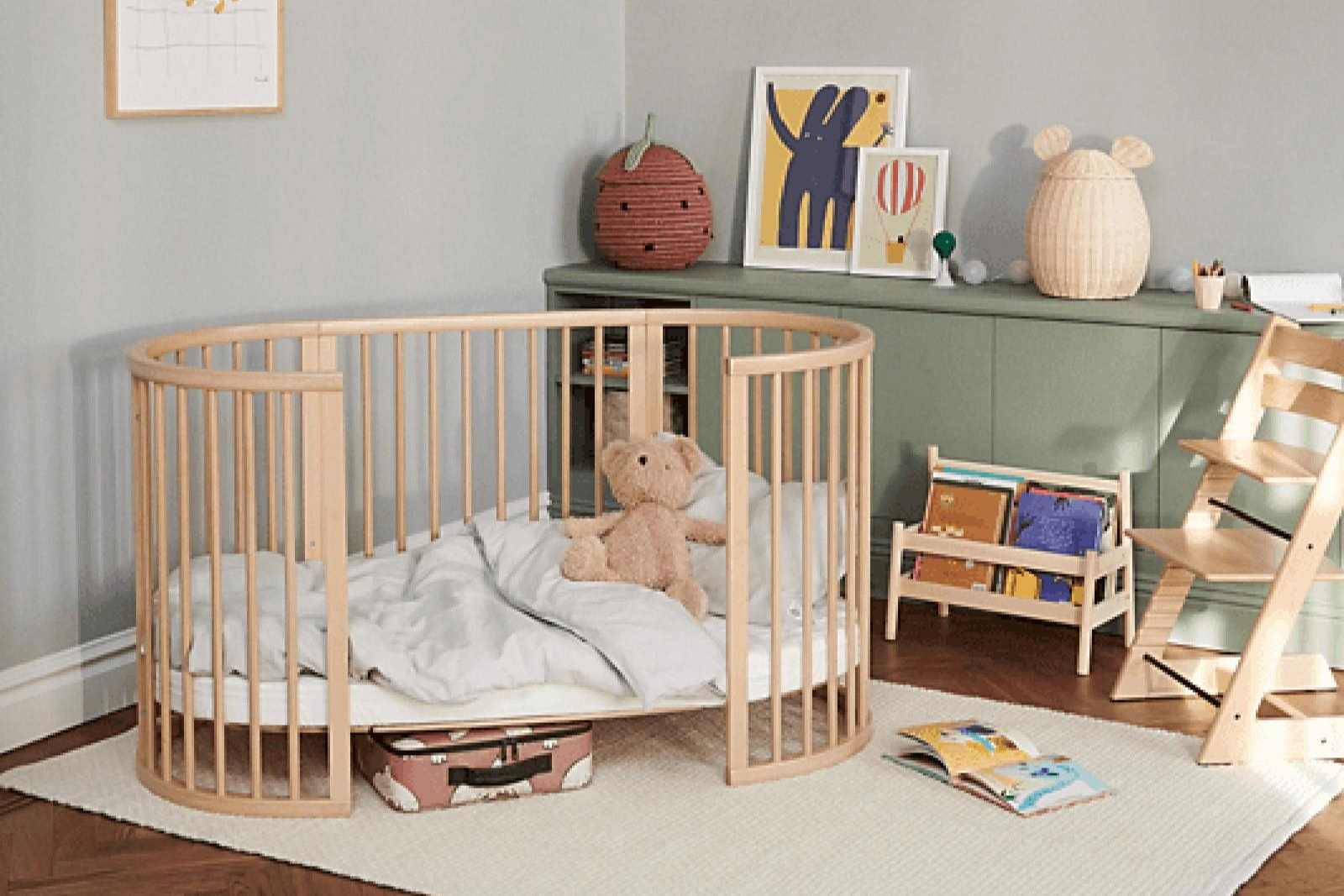 Modern nursery with oval wooden crib, teddy bear, colorful artwork, green dresser, and wooden bookshelf on white rug.
