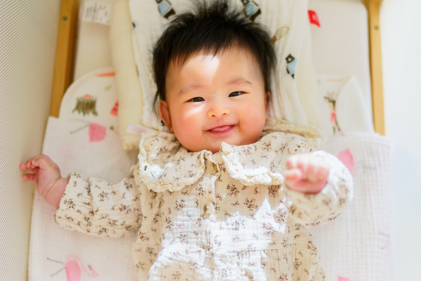 Smiling baby with dark hair lying in crib wearing floral patterned outfit on patterned bedding.